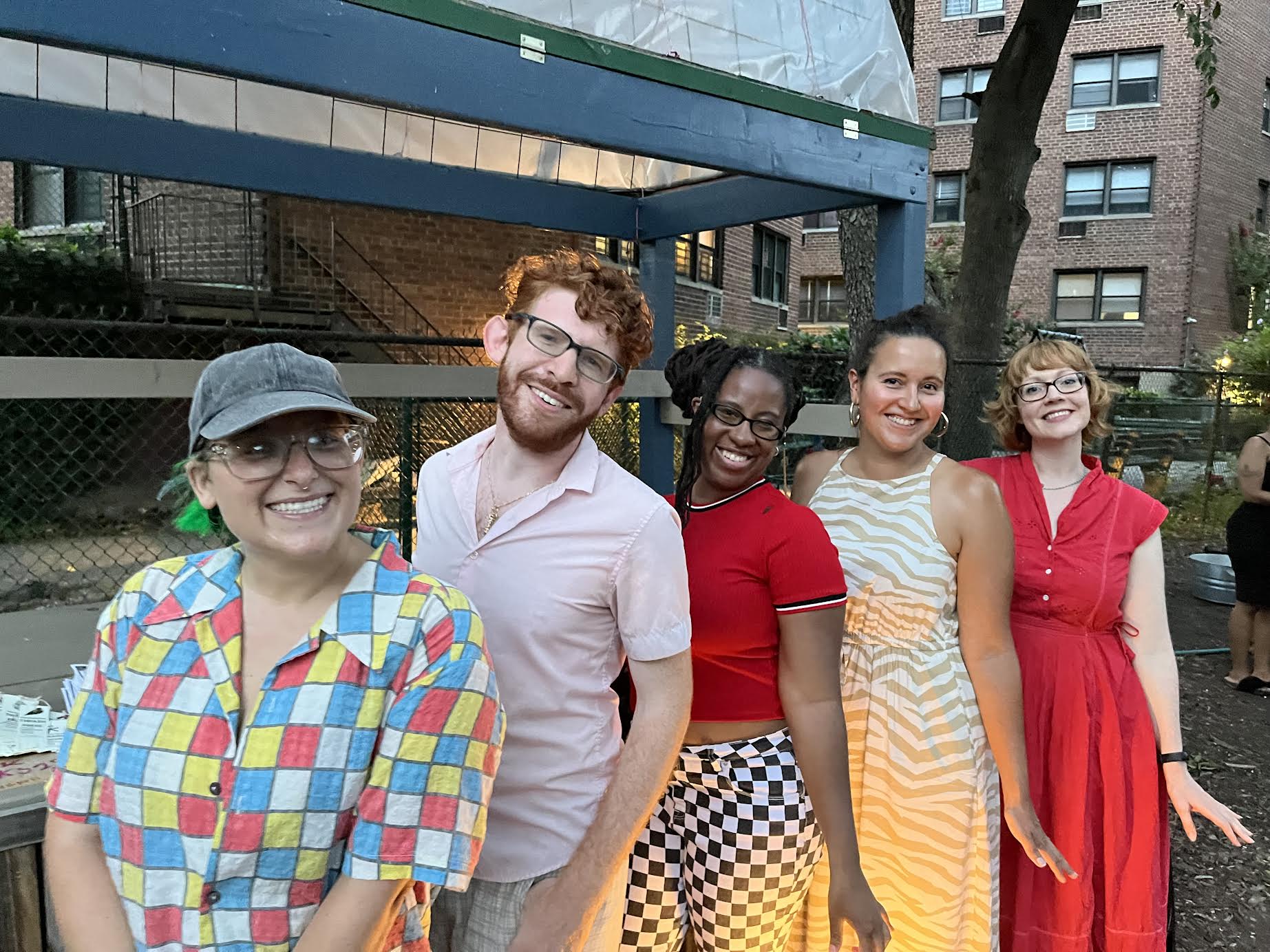 Five people in colorful outfits stand outdoors in front of a stage structure, smiling